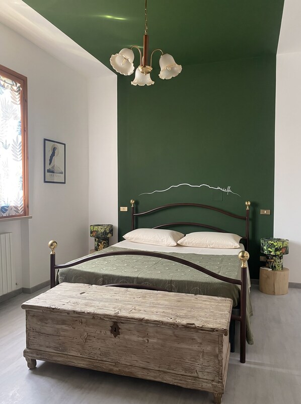 A bedroom with a bed, a wooden chest, a green wall, a chandelier, and a framed picture on the wall.