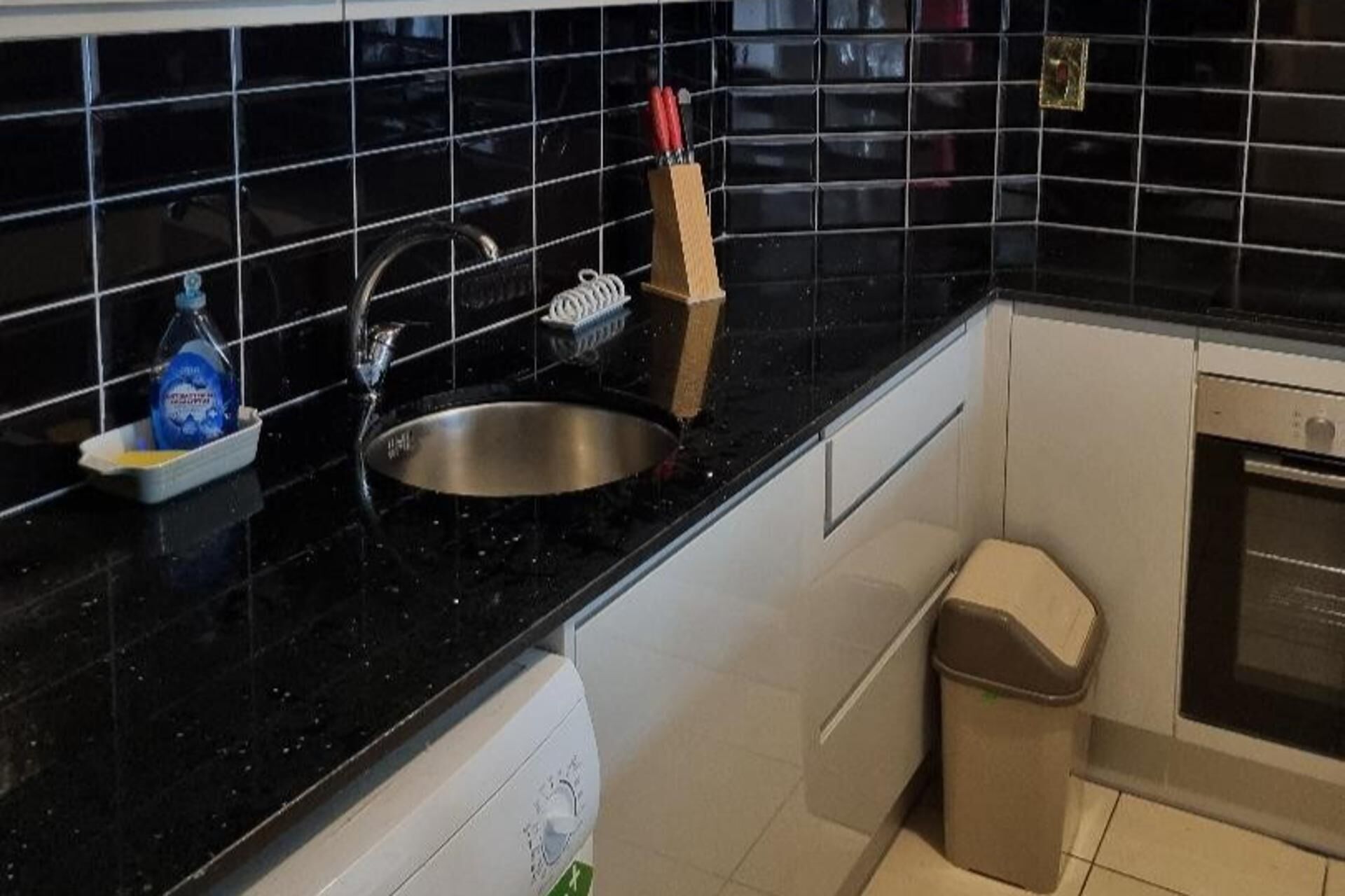 A kitchen with a black countertop, stainless steel sink, and a trash bin.