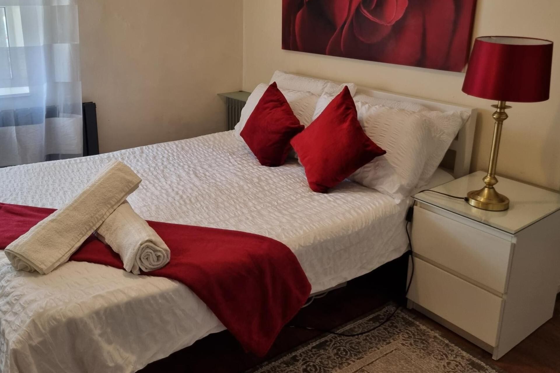 A neatly made bed with a white duvet, red pillows, and a rolled white towel. There is a red lamp on a bedside table and a painting on the wall.