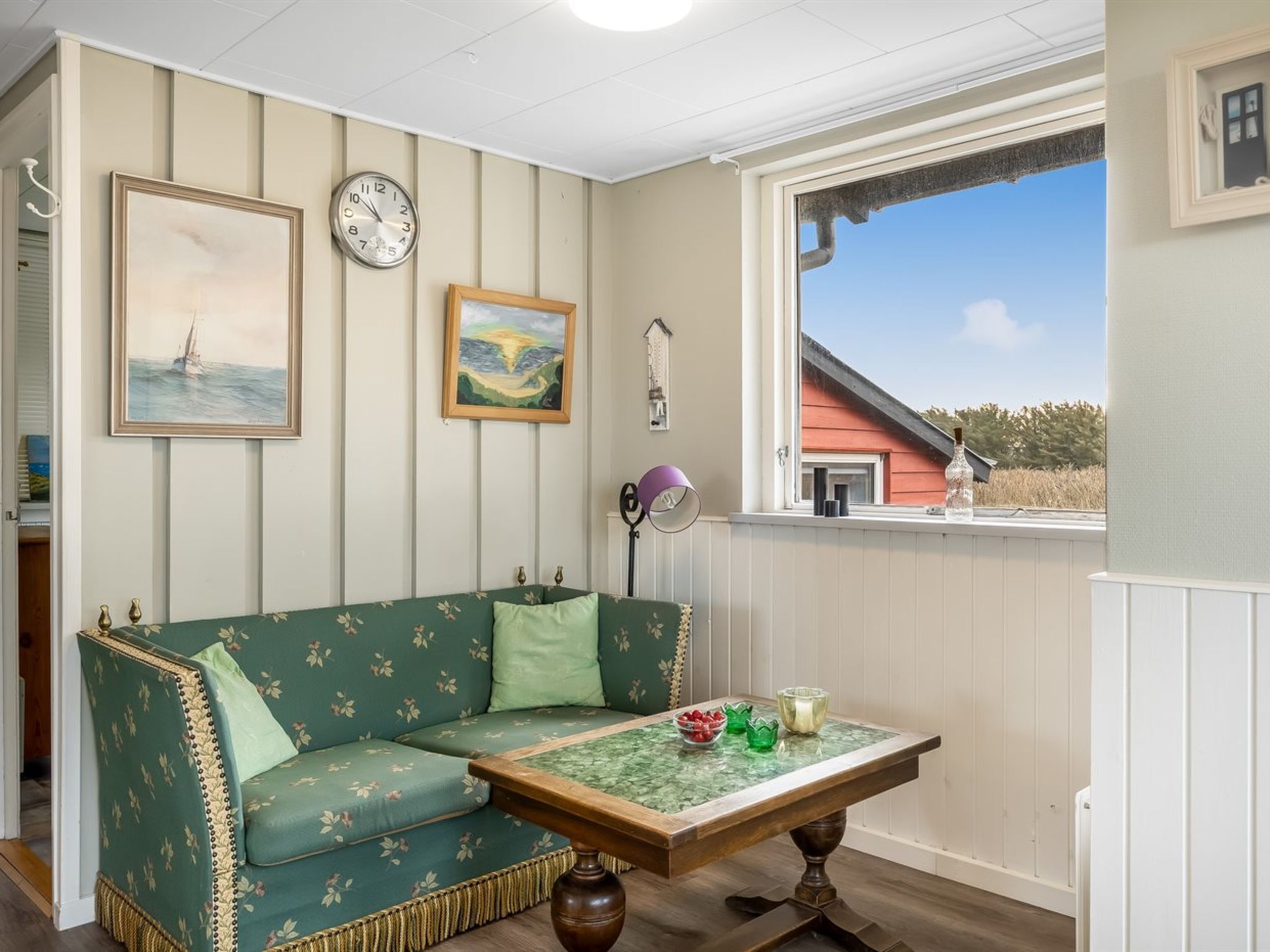 Una habitación con un sofá verde, una mesa de madera con cubierta de vidrio, un reloj y un ventanal con vista a un edificio rojo.