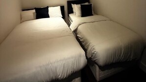Een kleine, zwak verlichte kamer met twee eenpersoonsbedden, elk met een zwart hoofdbord en witte beddengoed.