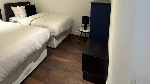 A hotel room with two beds, a small table, a lamp, and a dark-colored cabinet. - Cozy London Apartment With 2 Bedrooms (London)