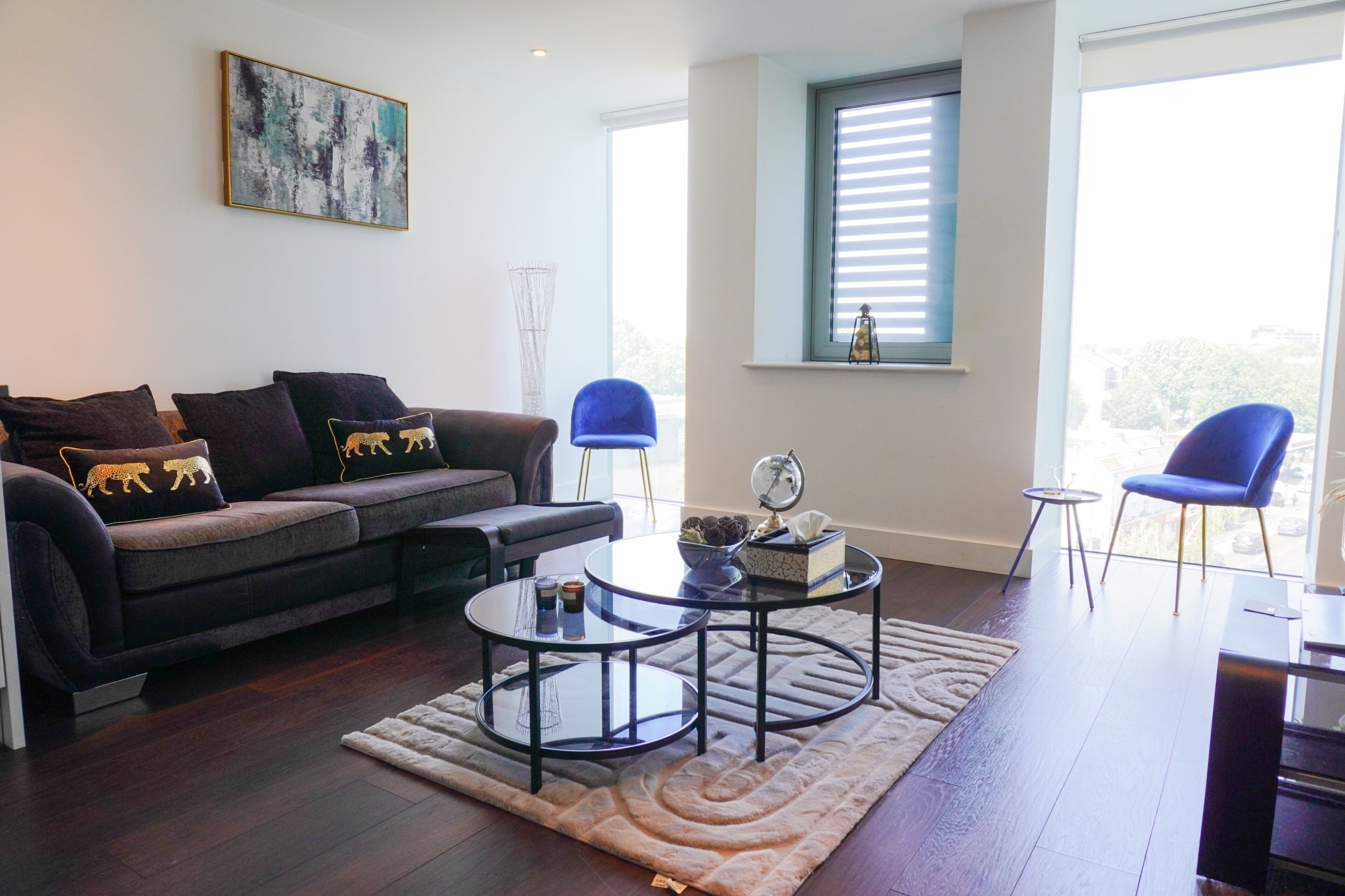 Een moderne woonkamer met een donkerblauwe bank, twee ronde salontafels, een blauwe fauteuil en een groot raam met zonwering.