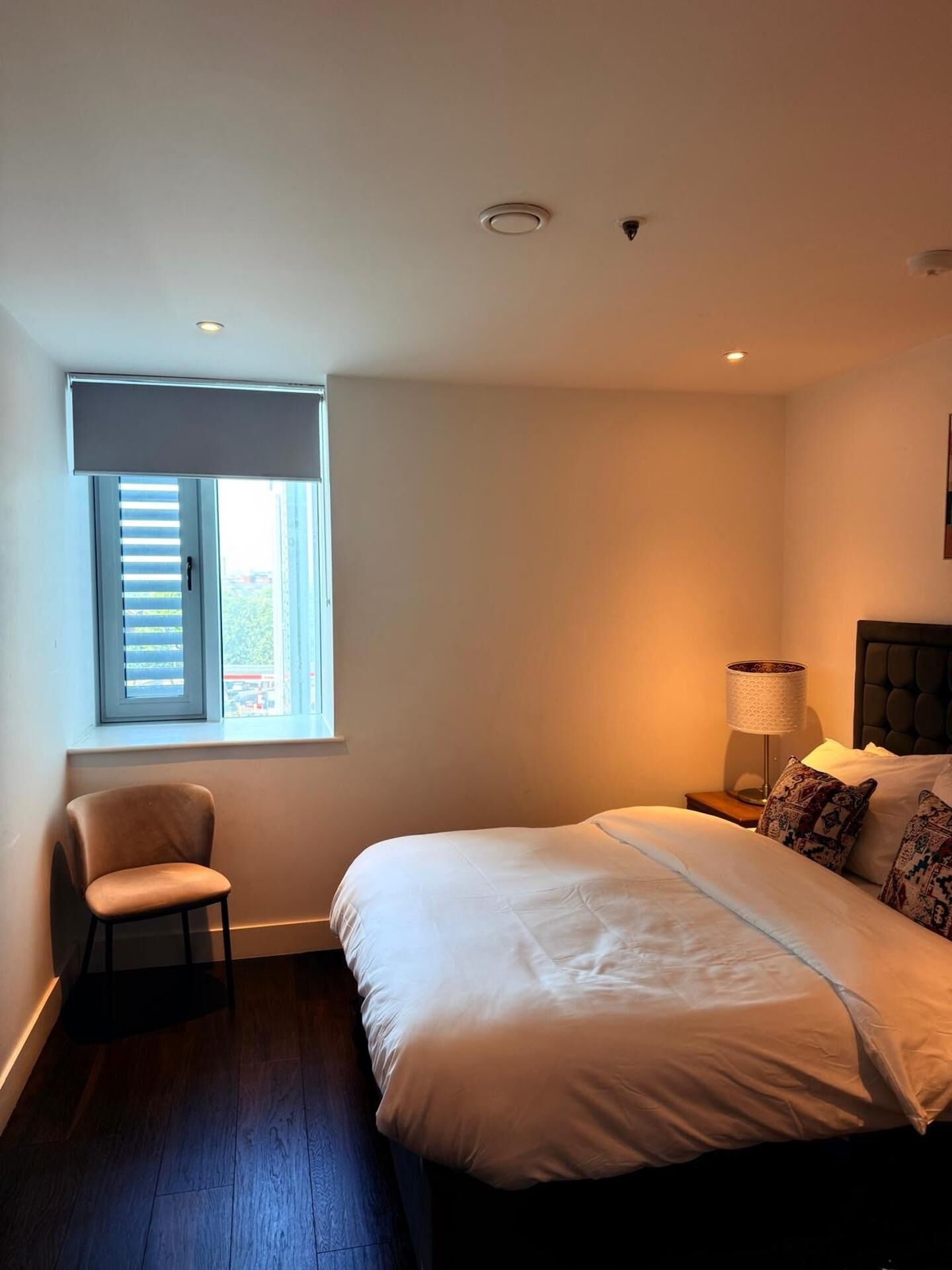 A bedroom with a bed, a chair, a nightstand, a lamp, and a window with blinds.