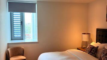 A bedroom with a bed, a chair, a nightstand, a lamp, and a window with blinds.