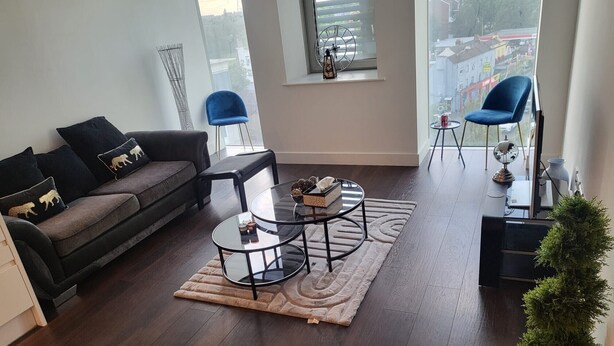 Een moderne woonkamer met een donker houten vloer, een grijze bank met decoratieve kussens, een ronde glazen salontafel en een uitzicht over de stad door grote ramen.