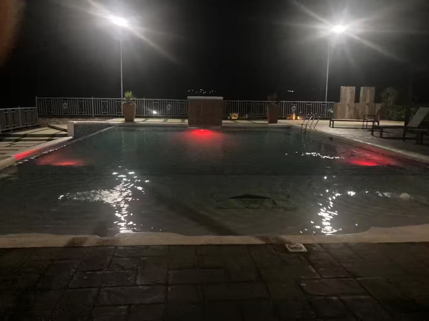 A nighttime swimming pool with two light poles, a wooden deck, and a metal railing.