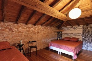 A bedroom with a stone wall, wooden ceiling beams, a bed with a pink bedspread, a bedside table, and a chair.