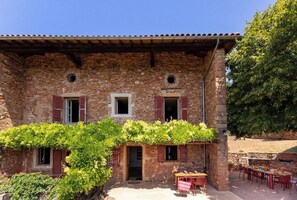 Ein rustikales Steinhaus mit überdachter Terrasse, Sitzgelegenheiten im Freien und einem Baum zur Rechten.