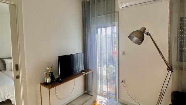 A modern living room with a flat-screen TV on a wooden stand, a floor lamp, a yellow patterned rug, and a sliding glass door leading to an outdoor area. - Casinha dos Carris- AL (Valença)