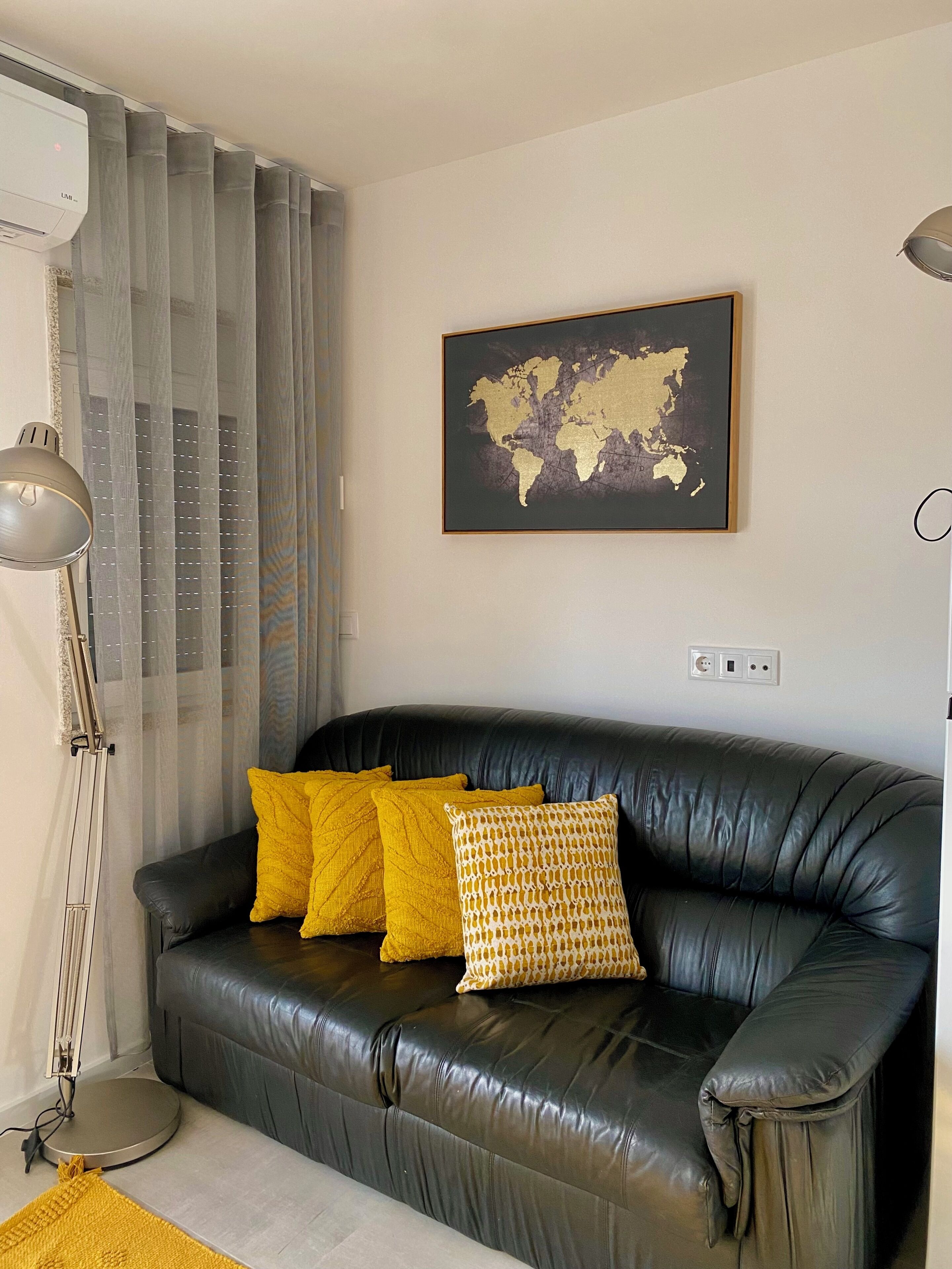 A black leather sofa with yellow and patterned cushions, a world map artwork, a floor lamp, and a radiator.
