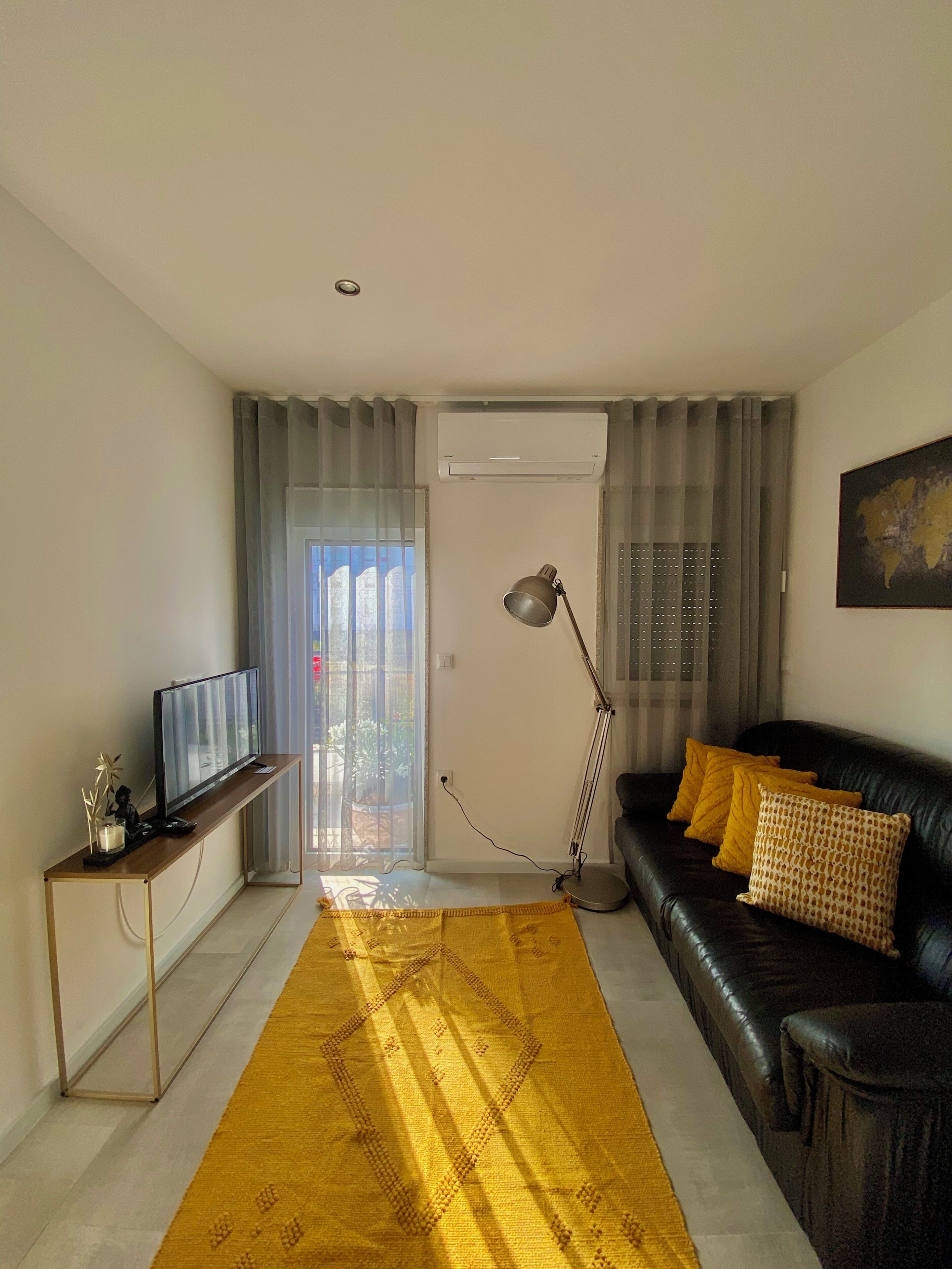 A living room with a TV on a stand, a sofa with cushions, a floor lamp, and a yellow rug.