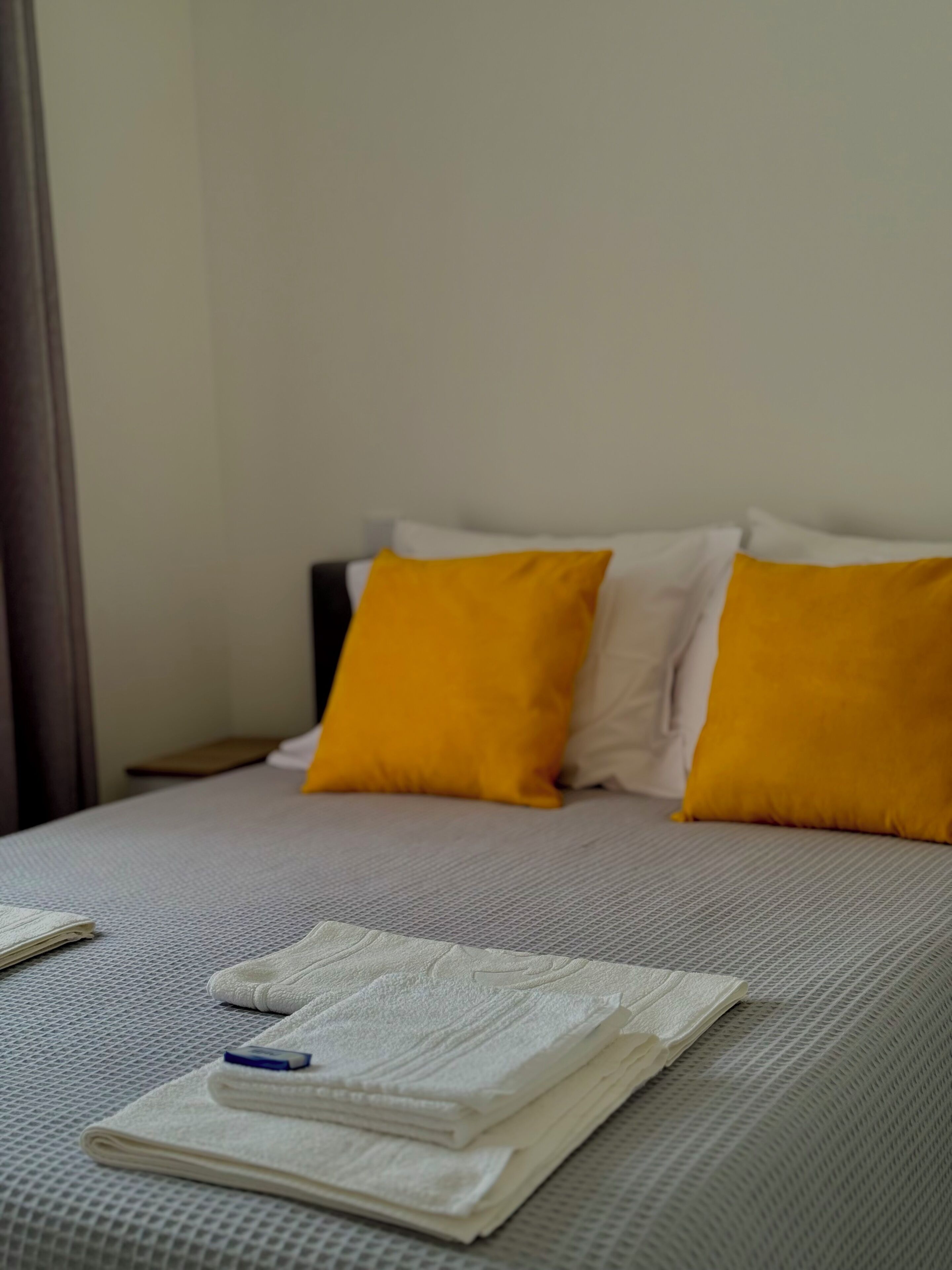 Ein ordentlich bezogenes Bett mit einer grauen, strukturierten Tagesdecke, zwei weißen und zwei leuchtend gelben Kissen.