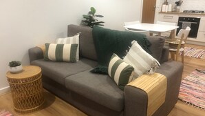 Ein Wohnzimmer mit grauem Sofa, gestreiften Kissen, einer grünen Decke, einem kleinen runden Weidenbeistelltisch und einem farbenfrohen Teppich.