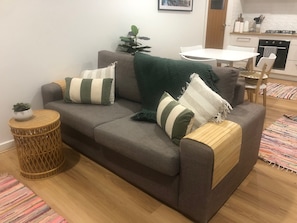 A living room with a grey sofa, striped pillows, a green throw blanket, a small round wicker side table, and a colorful woven rug.