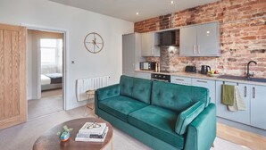 A modern living room with a green sofa, a wooden coffee table, and a kitchen with a brick wall and grey cabinets. - Umpires Watch - Cricket View - Free Parking (Scarborough)