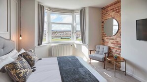 A hotel room with a large bed, a view through a window, a round mirror, a small table, and a chair. - Umpires Watch - Cricket View - Free Parking (Scarborough)