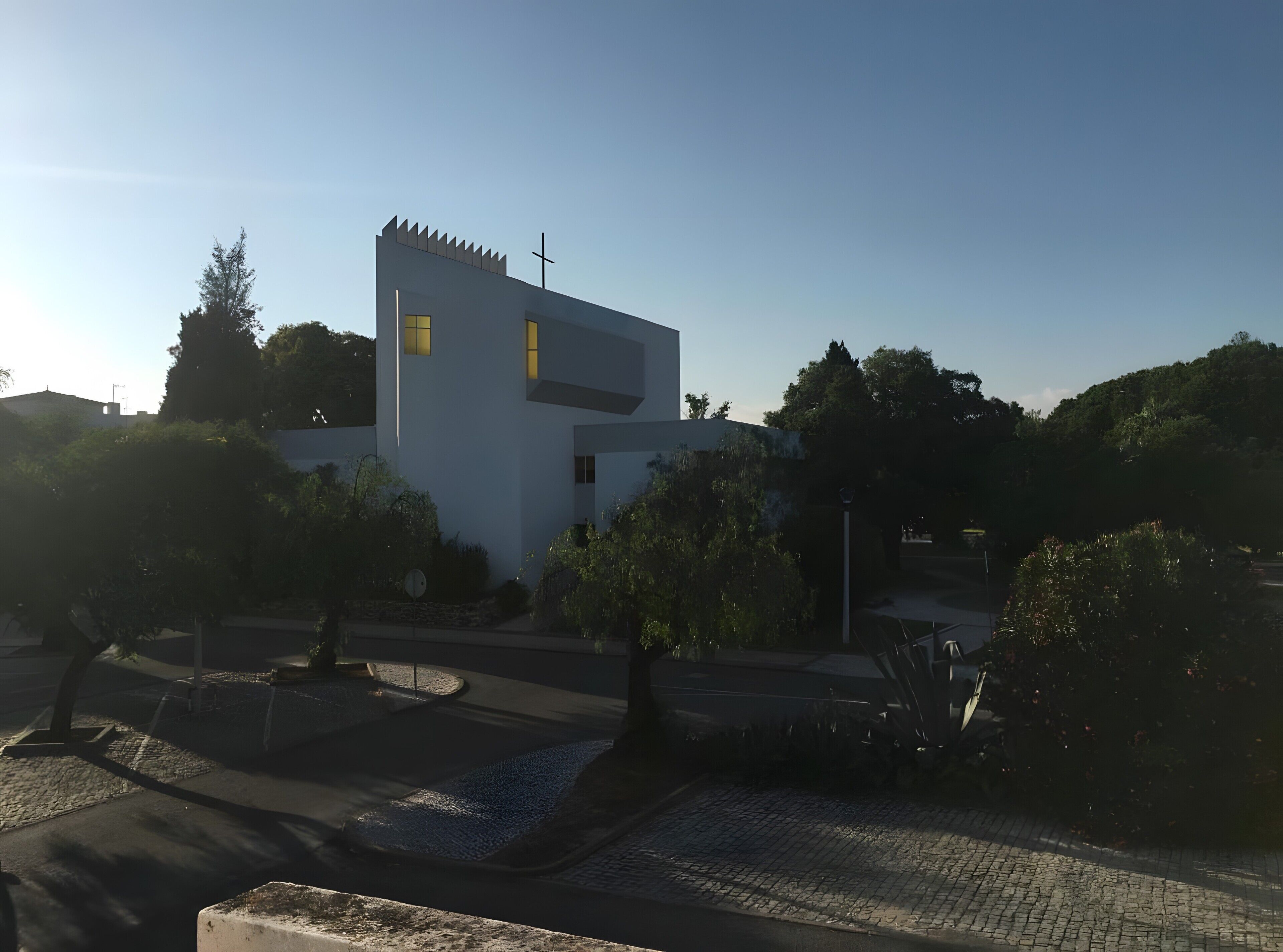 A modern building with a cross on top, surrounded by trees and a paved area.