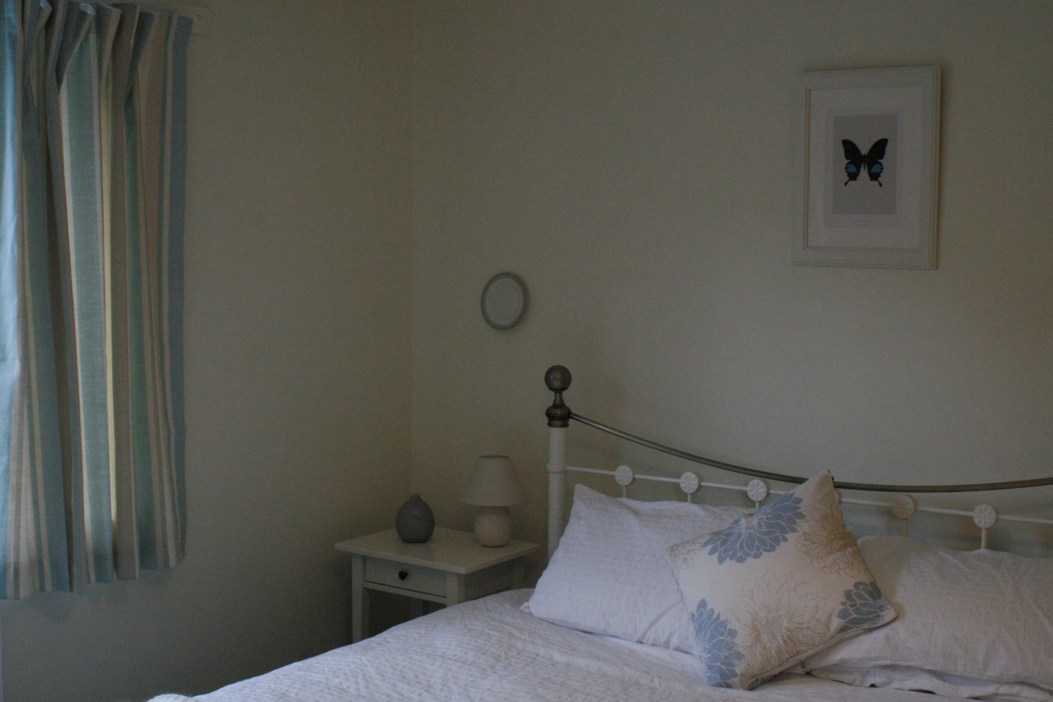A bedroom with a bed, pillows, a bedside table with a lamp, a wall clock, and a framed butterfly picture.