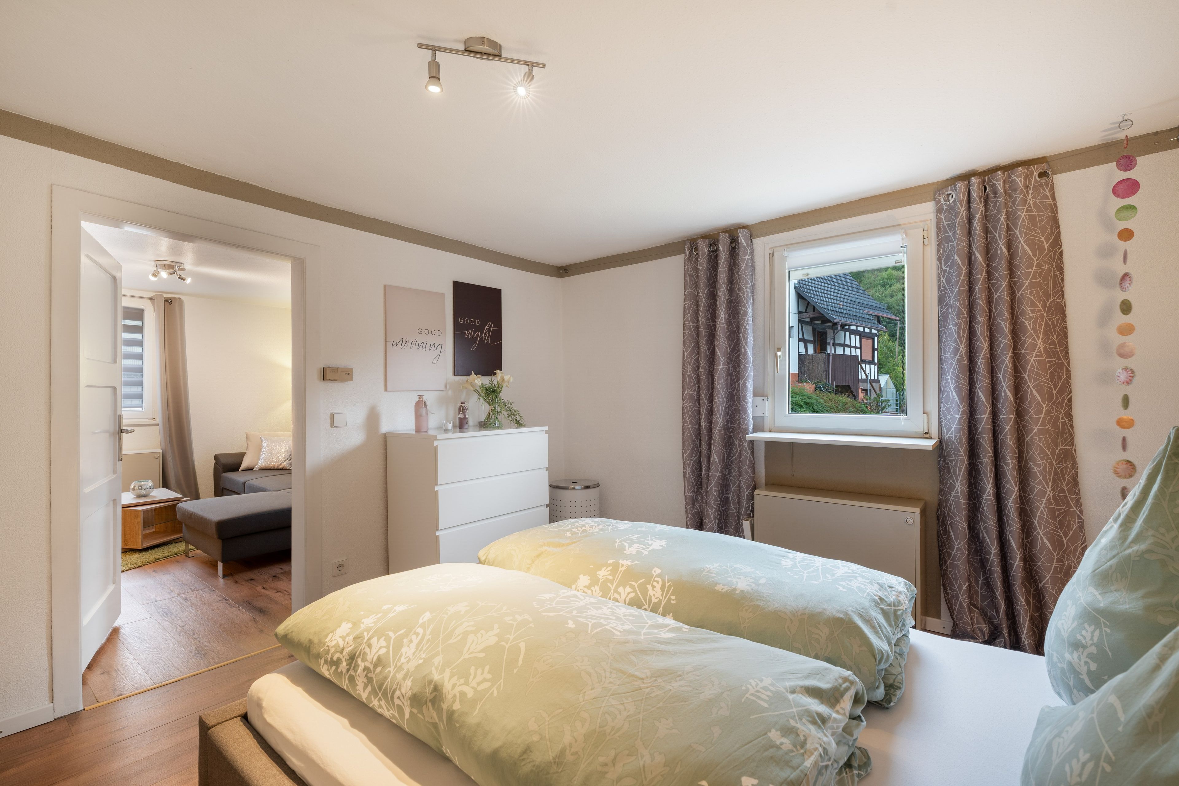 Ein Schlafzimmer mit Bett, Kommode, einem Fenster mit Vorhängen und Blick auf ein Haus außerhalb.