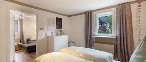 Un dormitorio con cama, cómoda, ventana con cortinas y vista a una casa.
