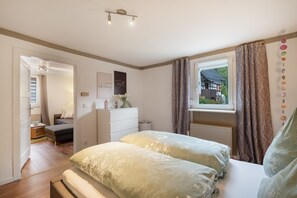 Ein Schlafzimmer mit Bett, Kommode, einem Fenster mit Vorhängen und Blick auf ein Haus außerhalb.