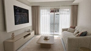 Ein modernes Wohnzimmer mit einem an der Wand befestigten Flachbildfernseher, einem beigen Sofa, einem Couchtisch und einer Glastür, die auf einen Balkon führt.