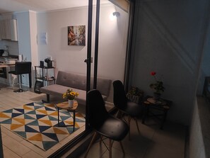Un salon avec un canapé, des fauteuils, une table basse, un tapis, un tableau et, au fond, une cuisine.