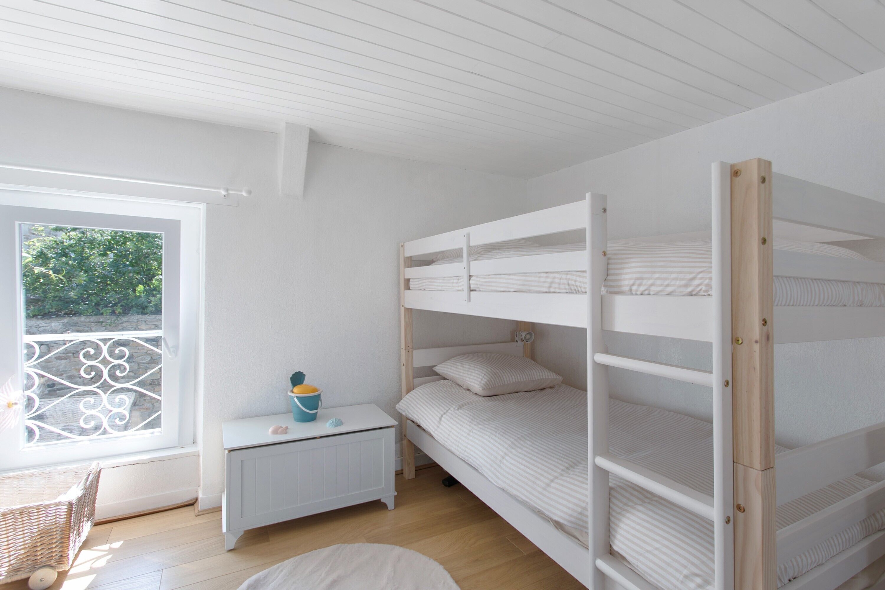 A bunk bed room with a window, a small white nightstand, and a wicker basket.