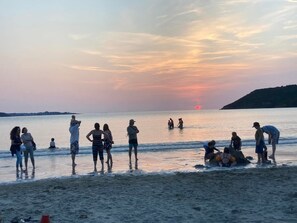 Une scène de plage où l’on voit des gens debout et jouant dans l’eau, un coucher de soleil en arrière-plan et une colline au loin.