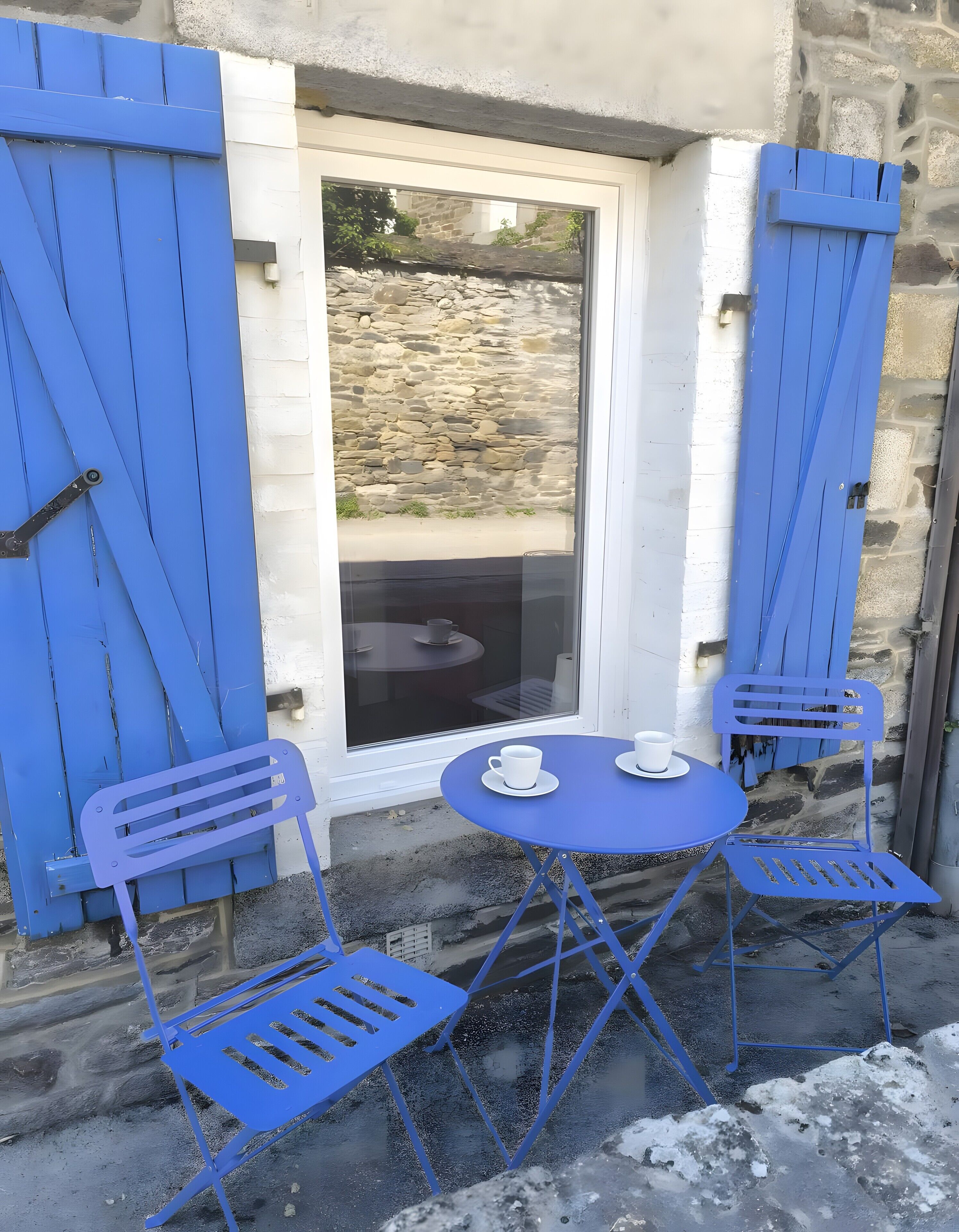 Ein kleiner Außenbereich mit Tisch und zwei Stühlen, ein Fenster mit blauen Rollläden und eine Steinsäule im Hintergrund.