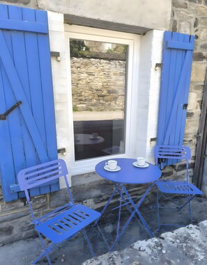 A small outdoor seating area with a table and two chairs, a window with blue shutters, and a stone wall in the background.