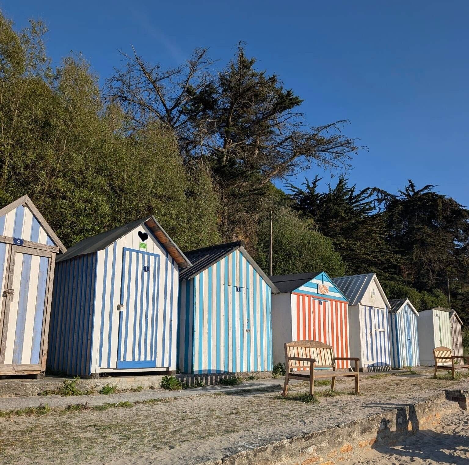 Une rangée de cabanes de plage aux façades rayées, un banc en bois et un ciel dégagé.