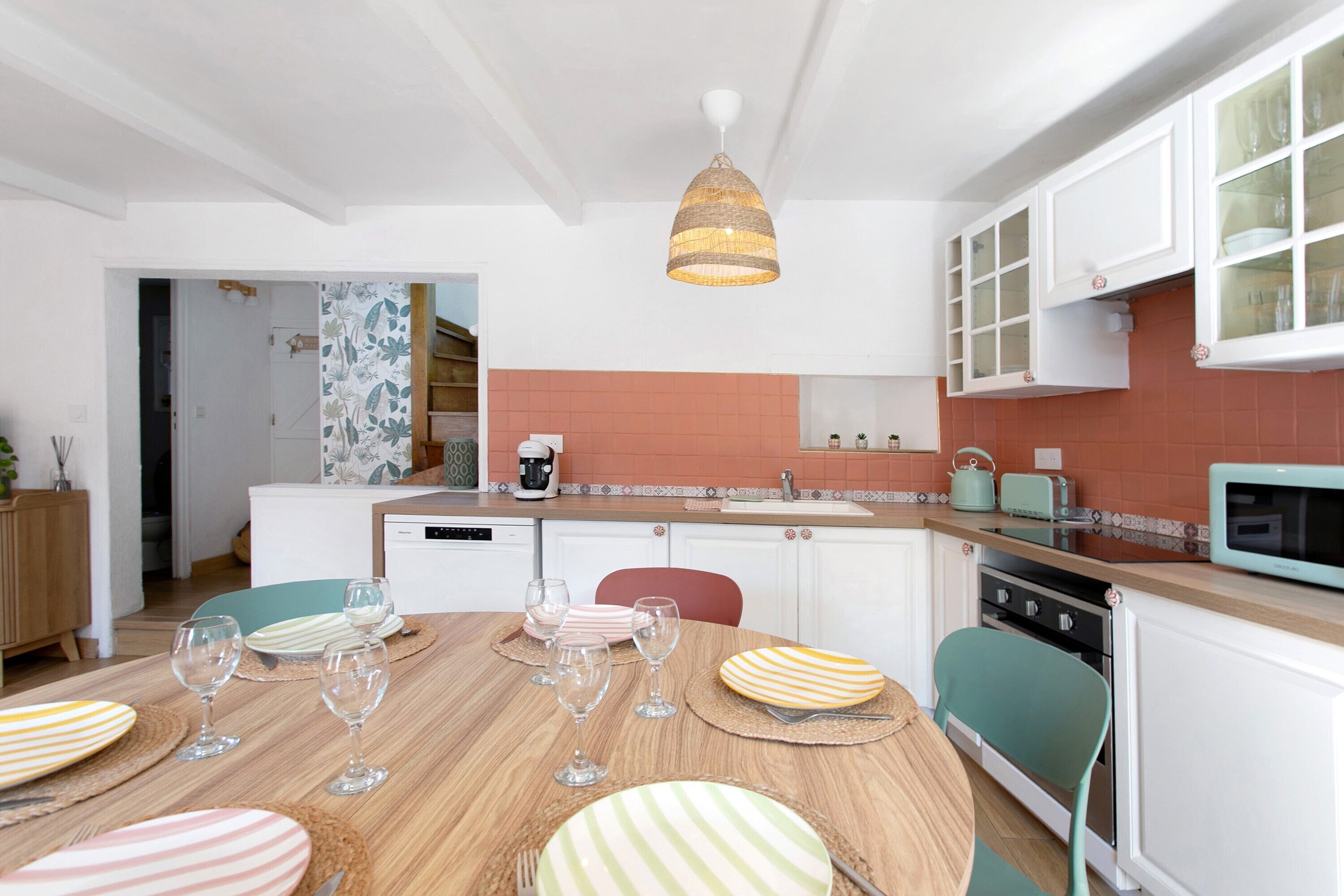 Une cuisine moderne avec des meubles blancs, une table à manger en bois mise avec des assiettes et des verres, et un luminaire suspendu.