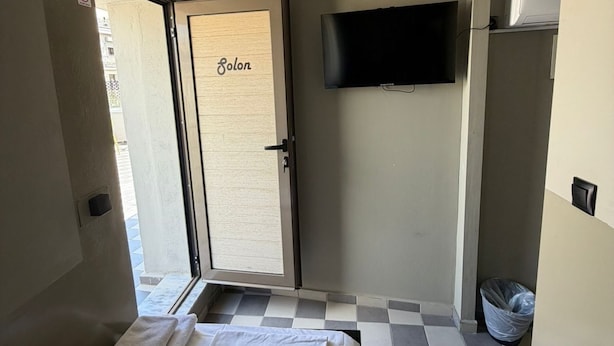 A hotel room with a bed, a TV mounted on the wall, a door labeled 'Solon,' and a checkered floor. - Petite rooftop room for 1person (ATHENS)