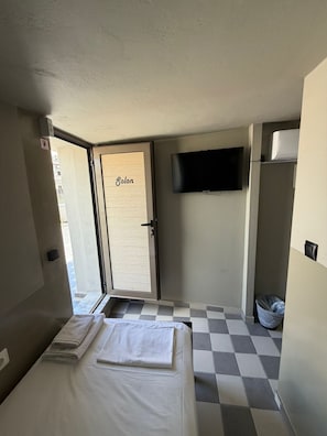A hotel room with a bed, a TV mounted on the wall, a door labeled 'Solon,' and a checkered floor. - Petite rooftop room for 1person (ATHENS)