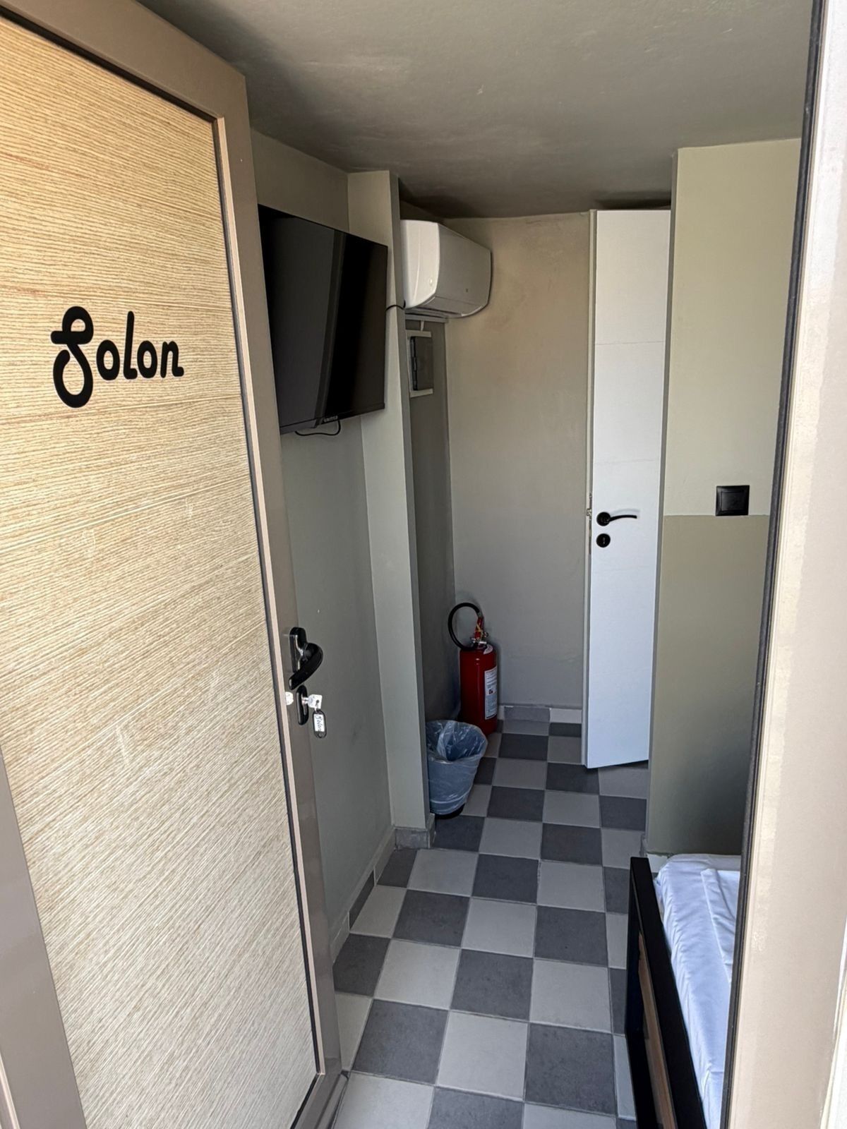 Ein kleiner Raum mit kariertem Boden, einem an der Wand montierten Fernseher, einem Feuerlöscher und einer Tür mit der Aufschrift '8olon'.