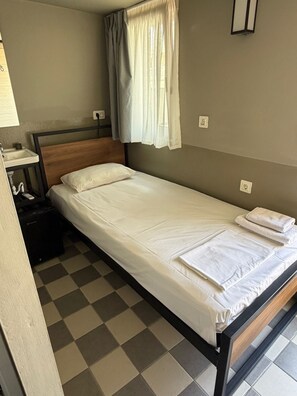 Ein Einzelbett mit weißer Bettwäsche, ein Holz-Kopfteil, ein kleiner Nachttisch mit Lampe und ein karierter Boden.