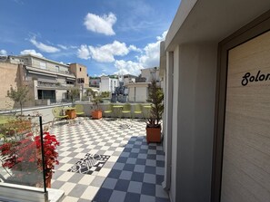 Eine Dachterrasse mit Schachbrettboden, grünen Stühlen, Blumentöpfen und Blick auf umliegende Gebäude.
