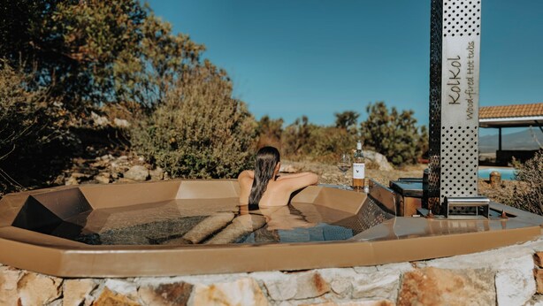 Une femme se détend dans un bain à remous, une bouteille de vin et un verre à la main, entourée d’un mur de pierres et de végétation.