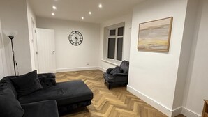A modern living room with a herringbone floor, a black sofa, a small armchair, a clock on the wall, and a window with a view.