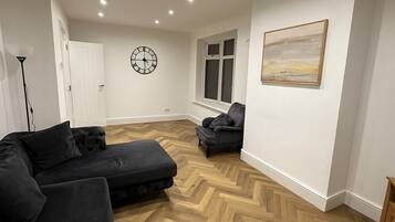 A modern living room with a herringbone floor, a black sofa, a small armchair, a clock on the wall, and a window with a view.
