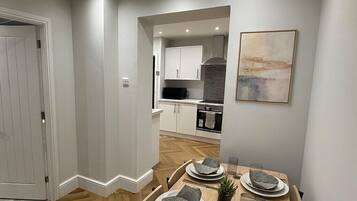 A modern dining area with a wooden table set for four, a kitchen in the background, and a painting on the wall.
