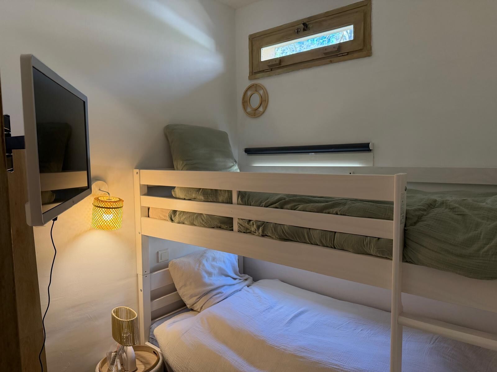 Uma cama de solteiro com uma televisão fixada na parede, uma janela com vista e uma mesinha com abajur.