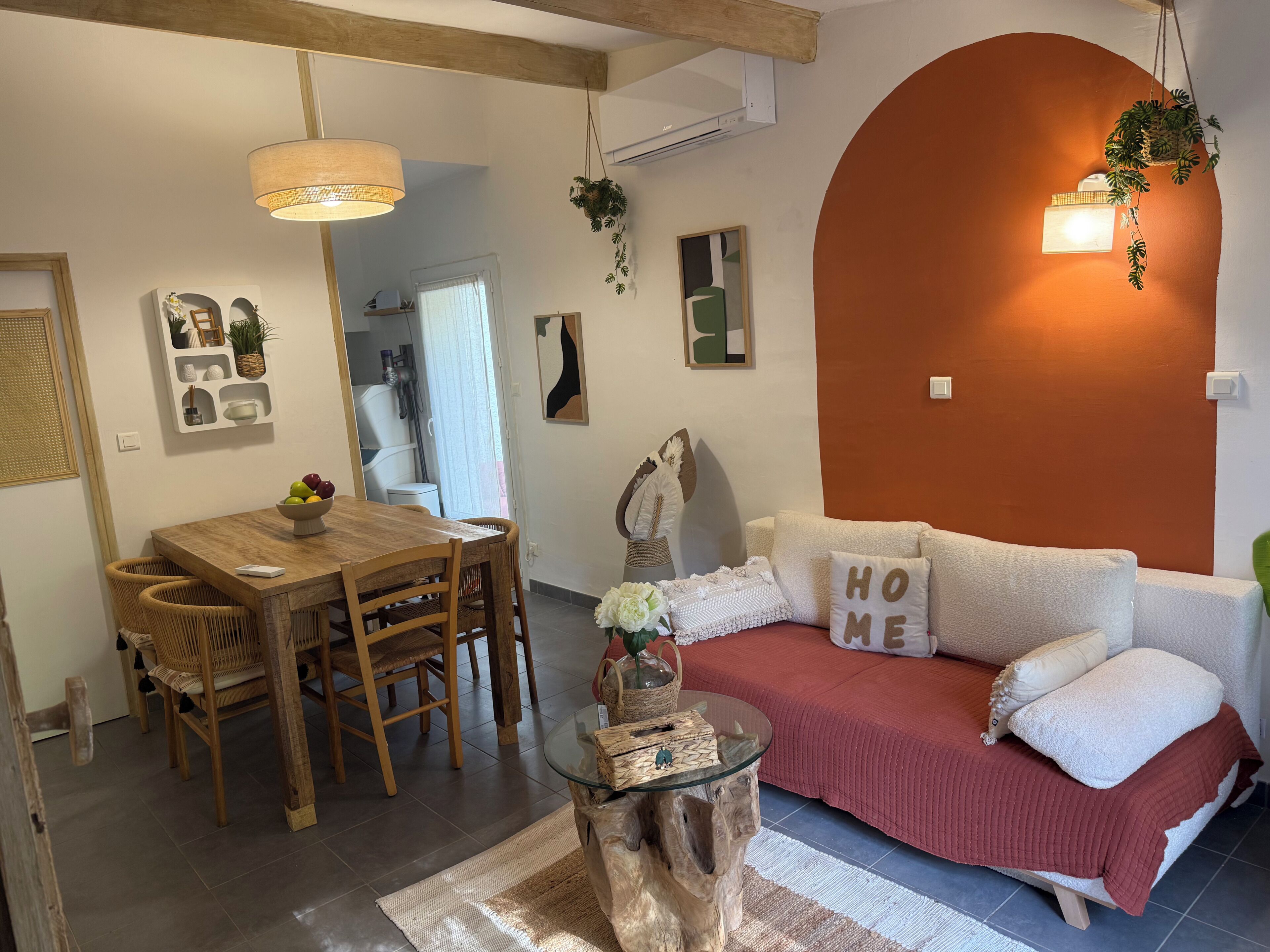 Un salon chaleureux avec une table à manger en bois, une table basse en verre, un canapé agrémenté d’un coussin orné du mot « MAISON » et un grand mur orange.