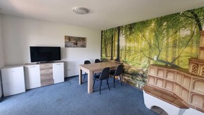A room with a dining table, chairs, a television, and a large forest mural on the wall. - Just like home, D3 OG? (Kreßberg)