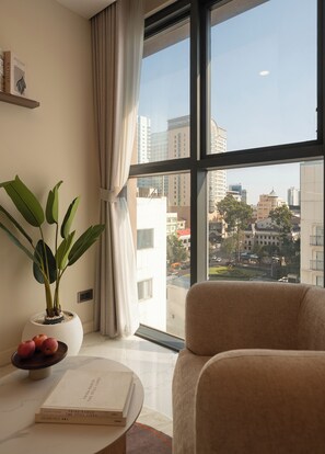 A modern living room with a large window, a potted plant, a sofa, and a table with books and apples. - Lux Studio in Madison by Maison De Gu (Thành phố Hồ Chí Minh)
