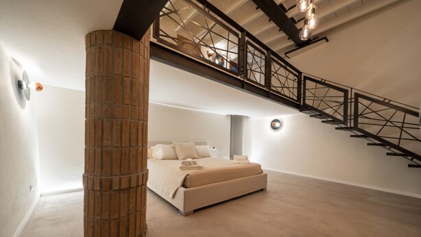 A bedroom with a bed, a large pillar, a staircase, and a ceiling light fixture. - In the heart of Olbia, the gateway to the famous Costa Smeralda, behind the facade of a historic building dating back to 1905, lies a place, renovated and designed by the renowned architect Lesuisse.This luxurious loft, with direct access from the street (Olbia)