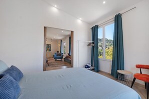 Ein Schlafzimmer mit einem großen Bett, einem Sessel, einem kleinen runden Tisch und Blick ins Freie durch ein Fenster.