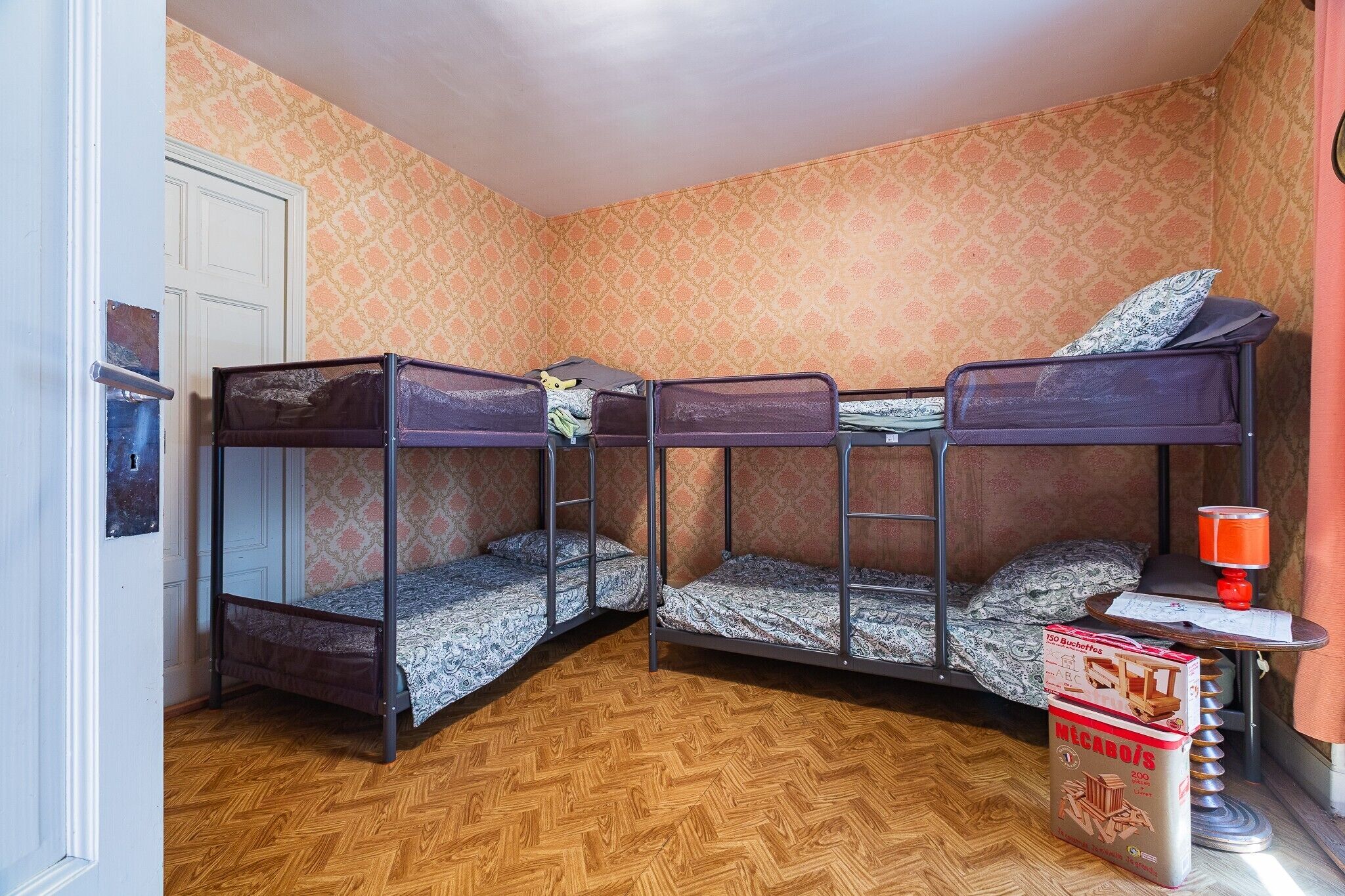 A room with three bunk beds, a wooden herringbone floor, a door, and a table with a lamp and a box of books.